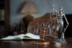 Themis and gavel on brown shining desk. Collection of legal books in the bookshelf.